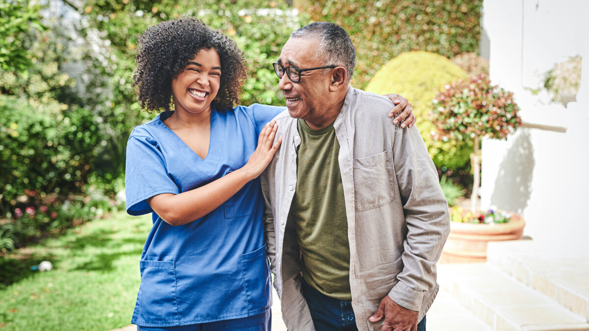 A healthcare professional in blue scrubs smiling and assisting an elderly individual with walking in a garden.