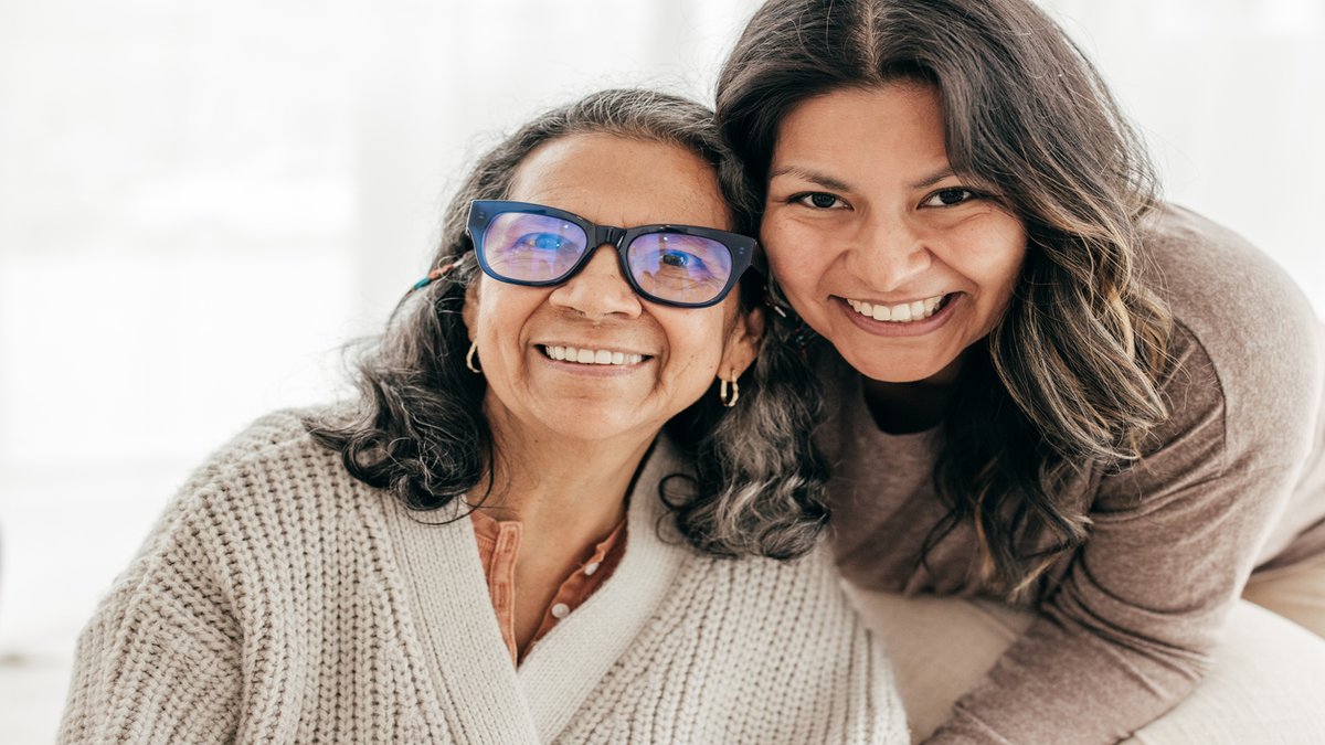 A smiling older woman wearing blue glasses and a knitted sweater, sitting closely with a younger woman who is leaning in and smiling warmly. Both appear happy and share a close bond, photographed in a bright and cozy setting.