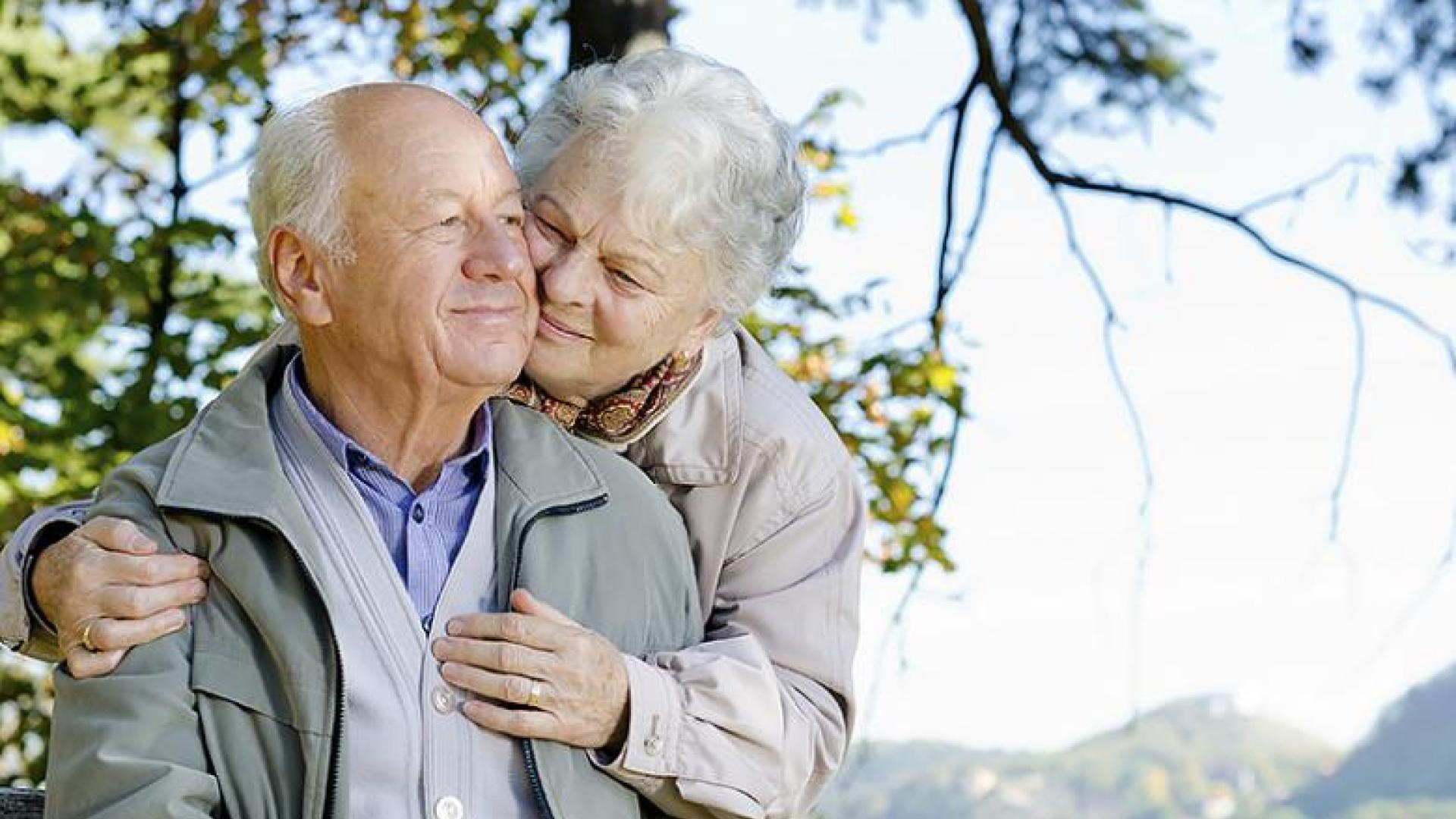 An old woman hugs an old man from slightly behind him and to his side. Their bodies are angled towards the camera but their faces are angled towards each other. There is a woods-like out-of-focus background.