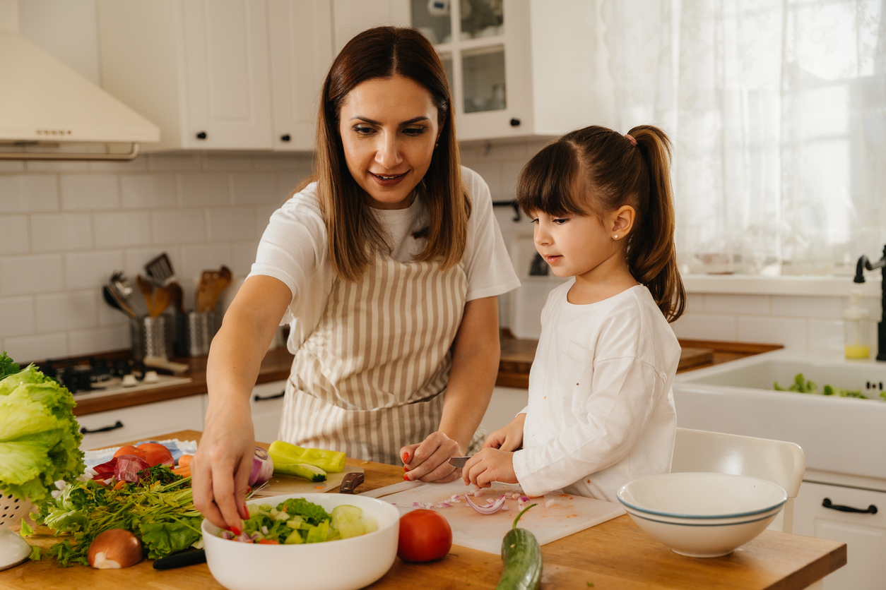 Cheerful young girl happily chopping vegetable with her mother. She appears curious and playful, enjoying the work. Her mother wearing a apron is also cutting vegetables while engaging with her in a lighthearted moment. The countertop is adorned with colorful fresh produce, including lettuce, tomatoes, and peppers.