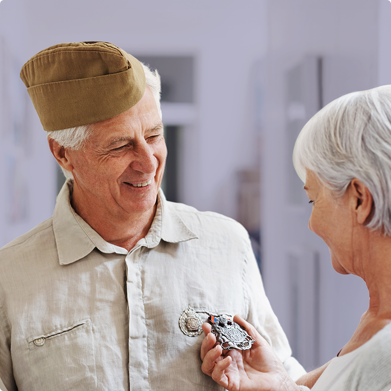 Elderly man in a vintage military-style hat smiling as a woman pins a medal onto his shirt in a warmly lit room.