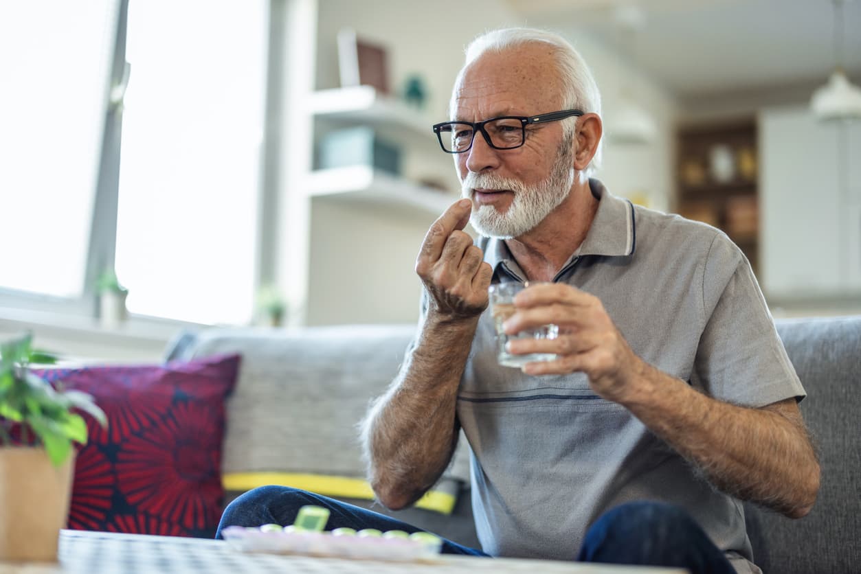 An older man sits on a couch with a glass of water in one hand and a pill pinched between the thumb and forefinger of the other, bringing the pill close to his mouth