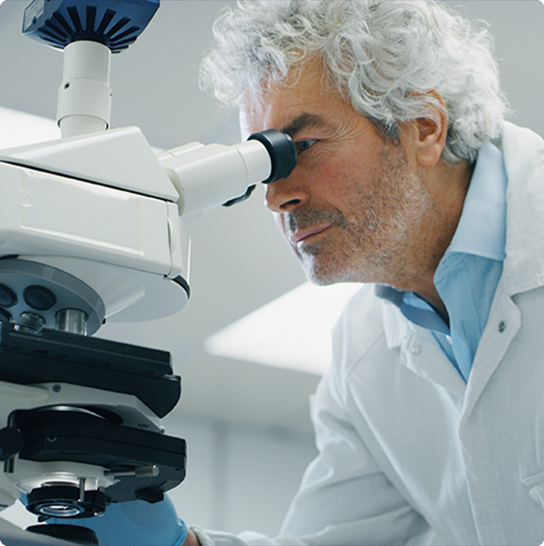 Scientist with gray hair wearing a lab coat examines a sample through a microscope in a laboratory.