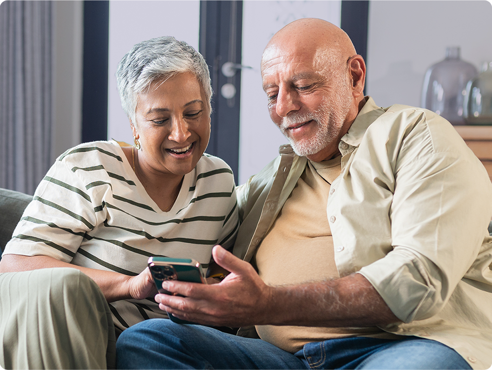 An older couple is sitting together on a couch, smiling and looking at a smartphone.