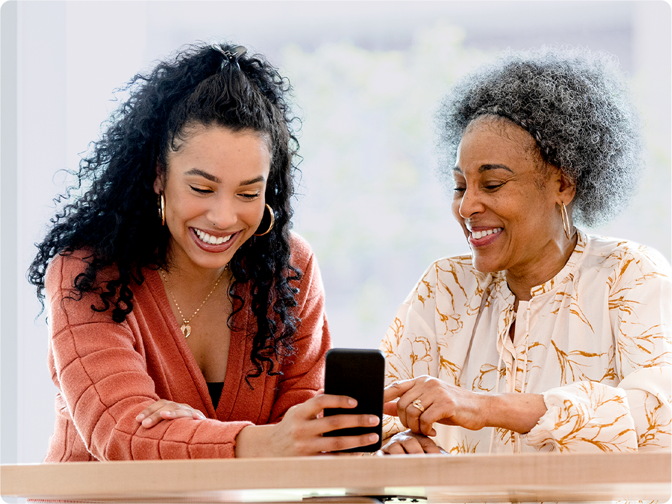 Two women, one younger and one older, are sitting together, smiling and looking at a smartphone.