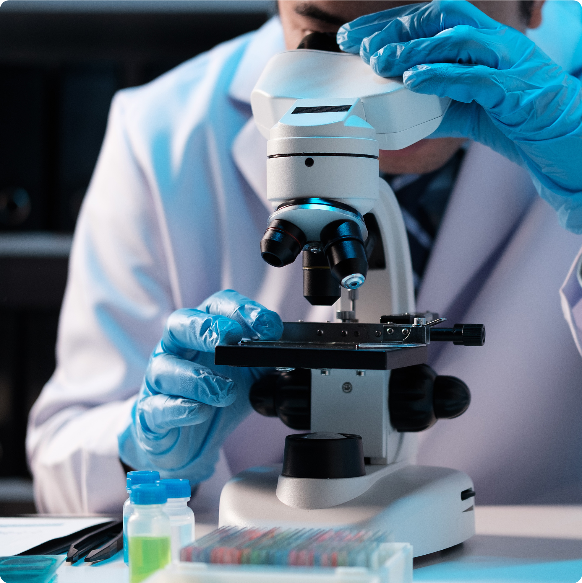 Scientist in a laboratory wearing a white coat and blue gloves, carefully examining a specimen through a microscope with sample vials and testing materials on the workbench.