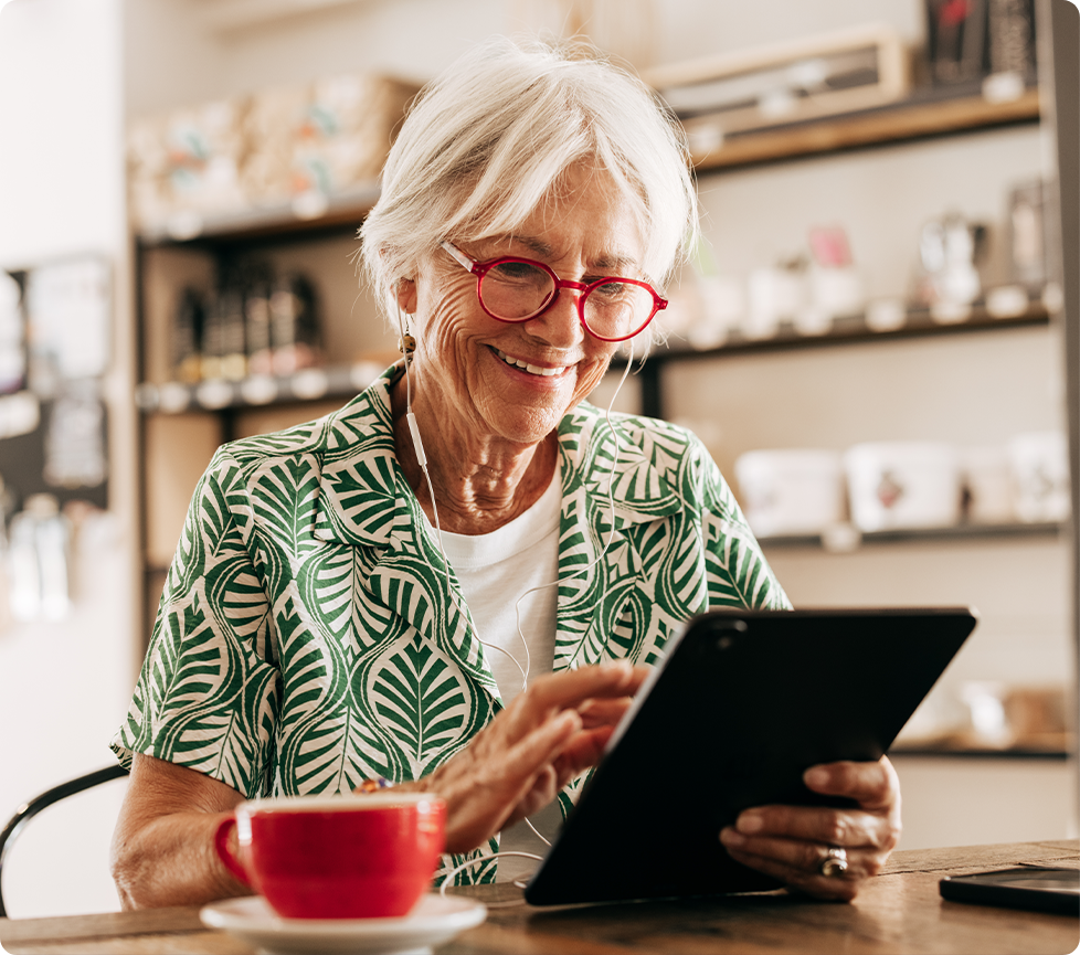 Smiling older adult wearing red glasses uses a tablet while sitting at a café table with a cup of tea and earbuds.