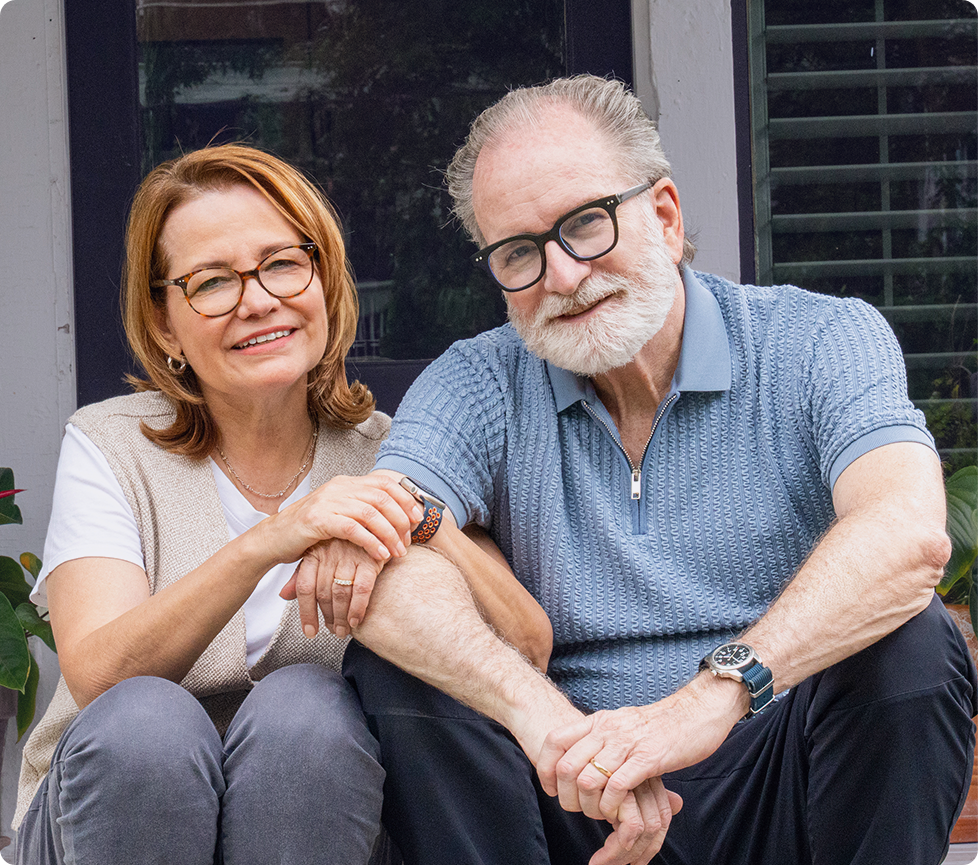 Smiling older couple wearing casual clothing and glasses, sitting closely together outdoors on the front steps of their home with their hands gently clasped, creating a warm and welcoming portrait.