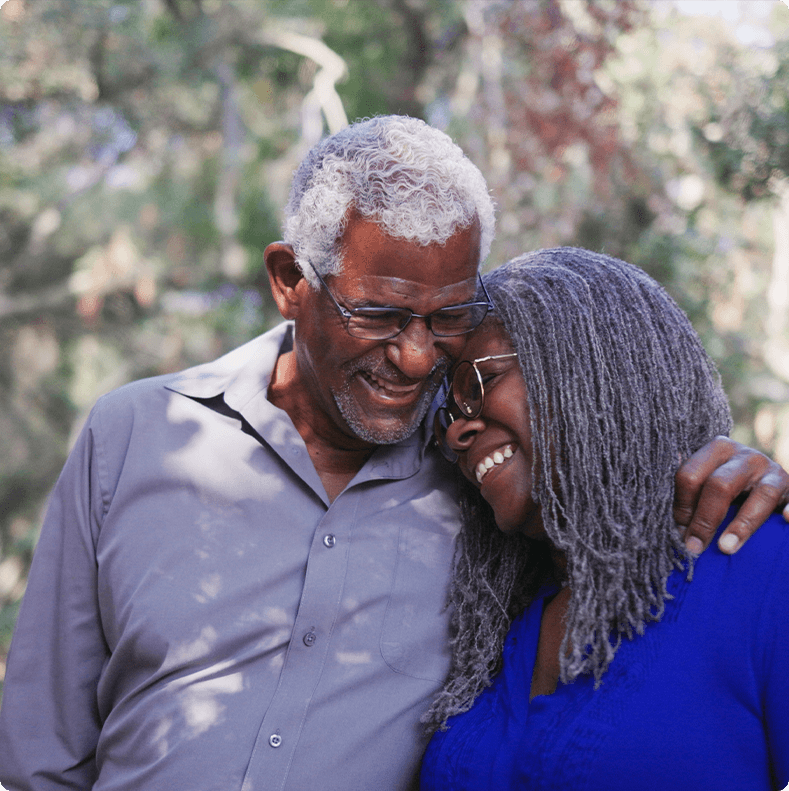 An older couple is standing close together outdoors, smiling and embracing.
