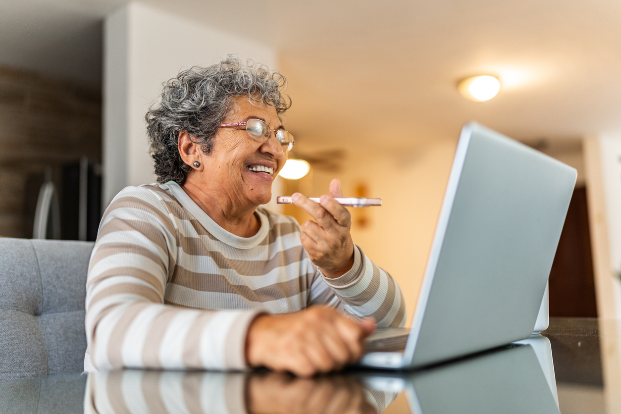 Senior woman using laptop and sending an audio message on mobile phone at home.